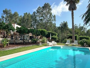 A swimming pool with white lounge chairs, surrounded by lush greenery and a stone wall.