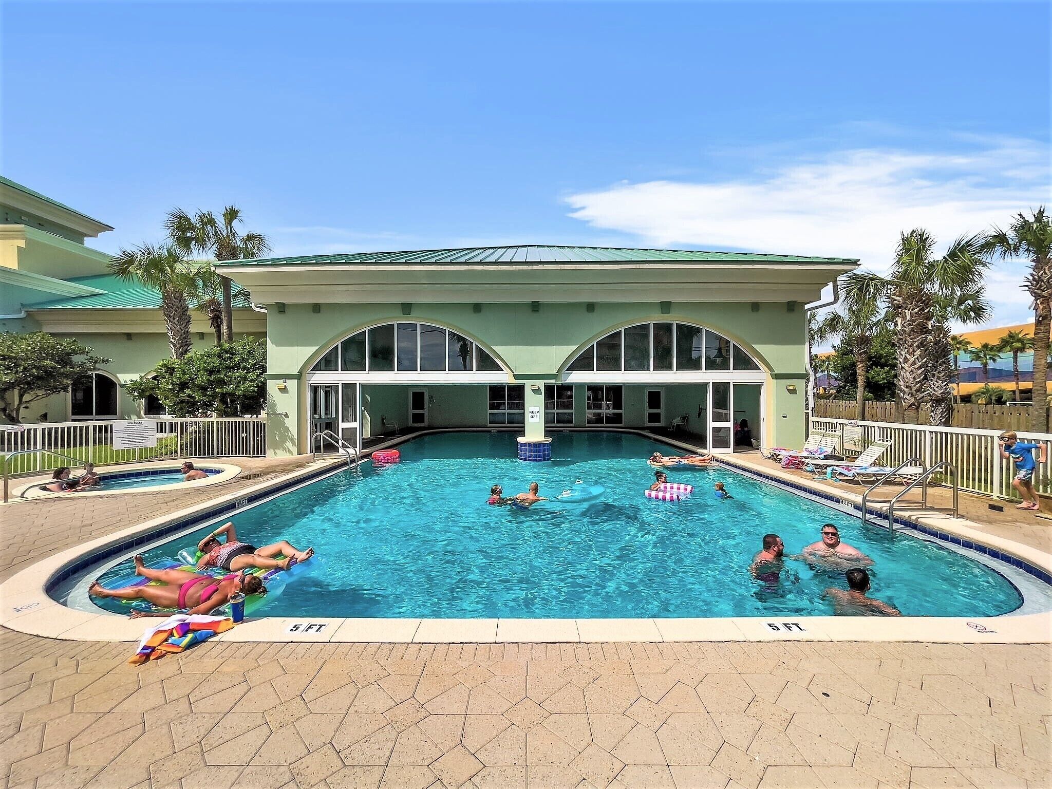 A swimming pool with multiple lanes, surrounded by palm trees and a building with arched windows.