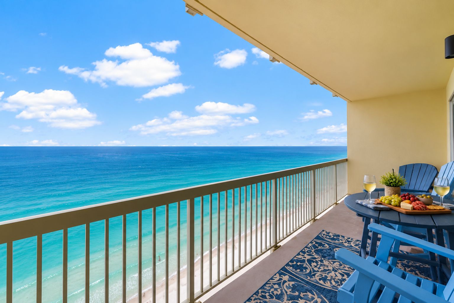 A balcony with a view of the ocean, a table with a fruit platter and wine glasses, and blue chairs.