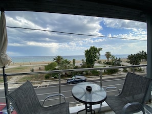 Un balcon avec une table en verre et deux chaises, donnant sur une plage bordée de palmiers et une route où circulent des voitures.