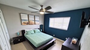 A bedroom with a bed, a ceiling fan, a window with blinds, and a bedside table with a lamp.