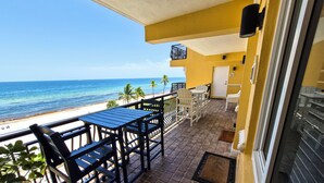 A balcony with a view of the beach and ocean, featuring a table and chairs, a potted plant, and a door with a number.