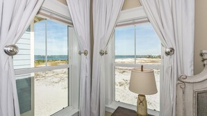 Habitación con vistas a la playa a través de amplios ventanales, cortinas blancas y una lámpara de mesa.