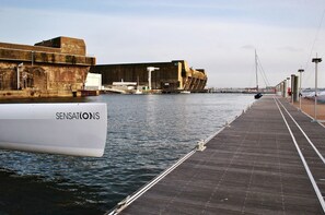 Eine Hafenbucht mit einem Boot namens 'SENSATIONS' und einer großen Industrieanlage im Hintergrund.