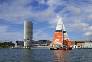 Een zeilboot met de tekst 'CITÉ DE VOILE' op het zeil, afgemeerd nabij een modern gebouw met een glazen gevel.