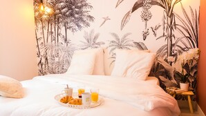 A neatly made bed with white linens, a tray of croissants, coffee cups, and orange juice, and a tropical-themed wall mural.