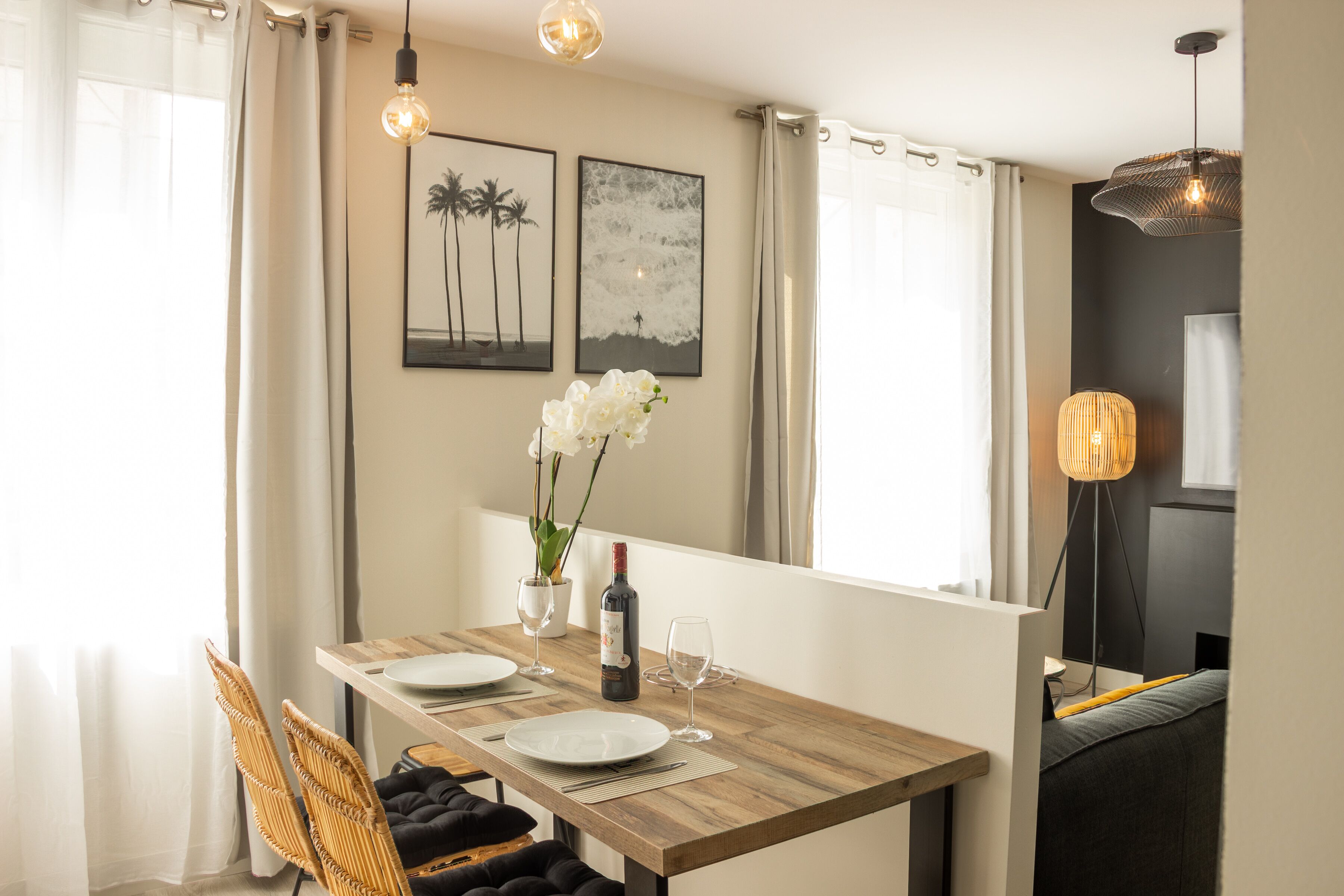 Un coin repas avec une table en bois, des assiettes blanches et une bouteille de vin. Deux tableaux encadrés ornent le mur, ainsi qu’un vase rempli de fleurs blanches et un lampadaire.