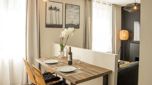A dining area with a wooden table, white plates, and a bottle of wine. There are two framed pictures on the wall, a vase with white flowers, and a floor lamp.