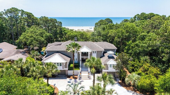 A large, multi-story house with a central entrance, surrounded by lush greenery and a view of the ocean.