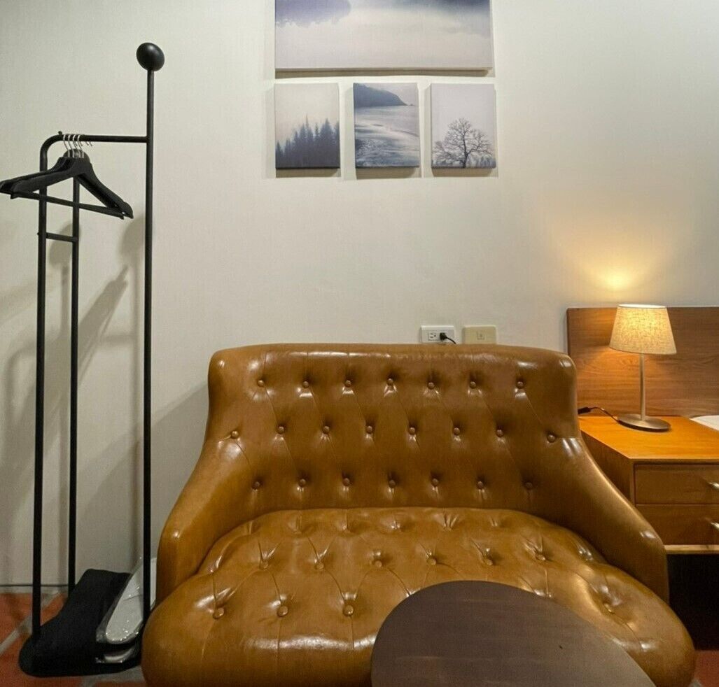 A brown tufted armchair in a room with a wooden nightstand, a lamp, and a coat rack with a single hanger.