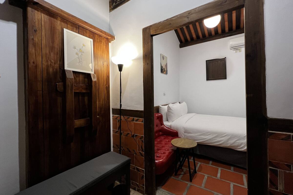 A small, cozy room with a bed, a bench, a stool, and a wall-mounted lamp.