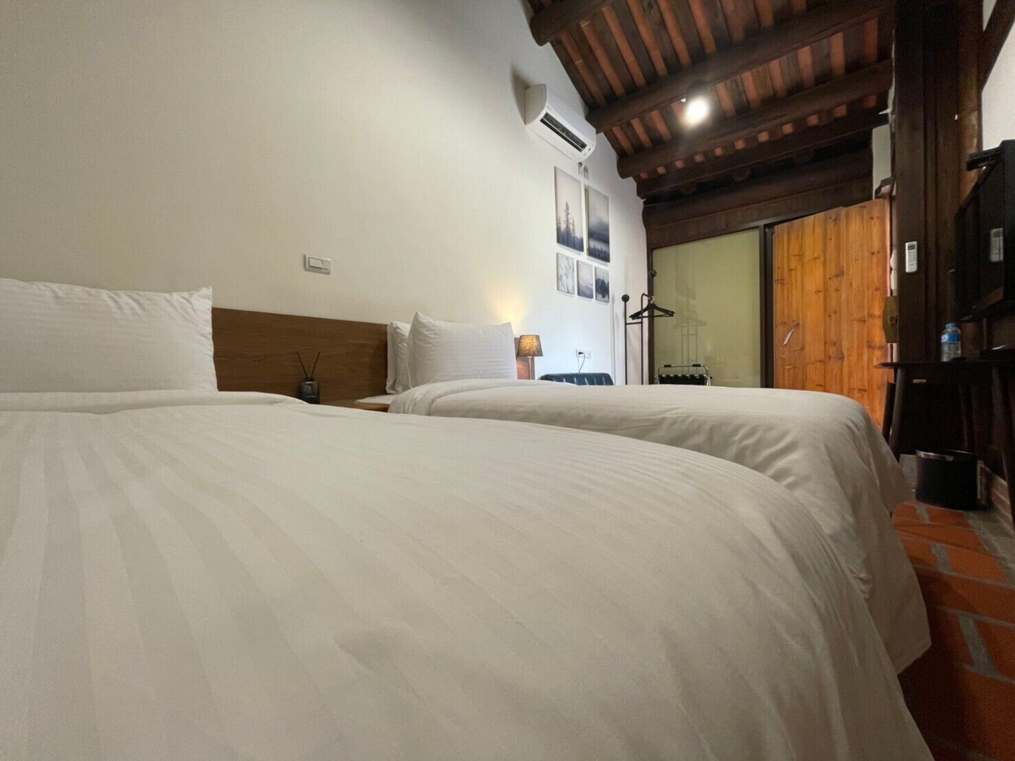 Habitación de hotel con dos camas, vigas de madera en el techo, aire acondicionado montado en la pared y una ventana con vistas al exterior.
