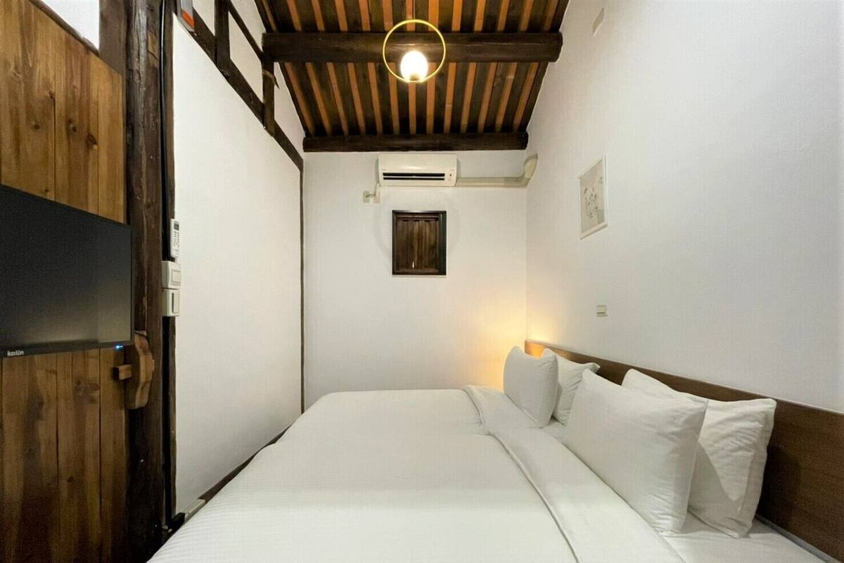 A bedroom with a wooden ceiling, a bed with white bedding, a wall-mounted air conditioner, and a small window.