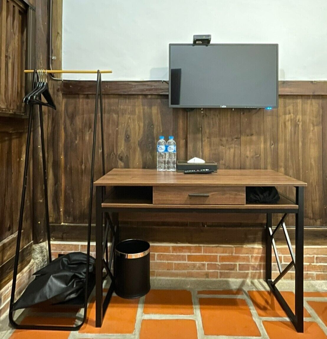 A wooden desk with a flat-screen TV, two water bottles, a black bag, and a trash can.