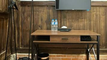 A wooden desk with a flat-screen TV, two water bottles, a black bag, and a trash can.