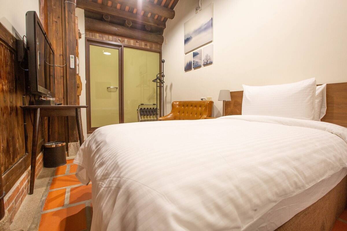 A bedroom with a large bed, wooden headboard, a television mounted on the wall, and a door leading to a bathroom.