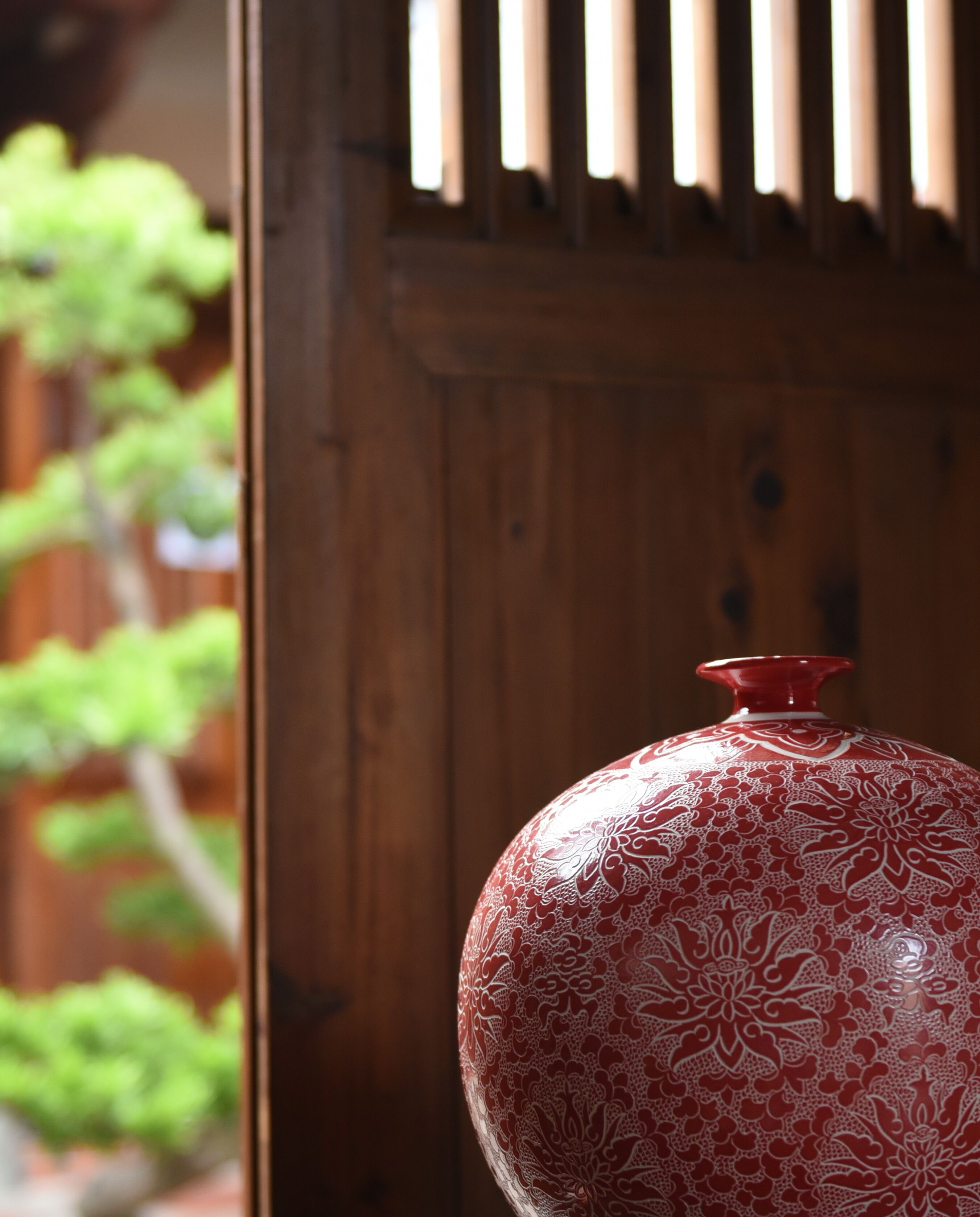 A traditional Japanese room with a wooden door, a red and white patterned vase, and a green plant in the background.