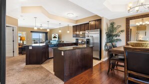 A spacious kitchen with dark wood cabinets, a central island, stainless steel appliances, pendant lighting, and a dining area with a large mirror and chandelier. - Branson Getaway • Sunroom + Balcony + Stunning Table Rock Lake View (Branson)