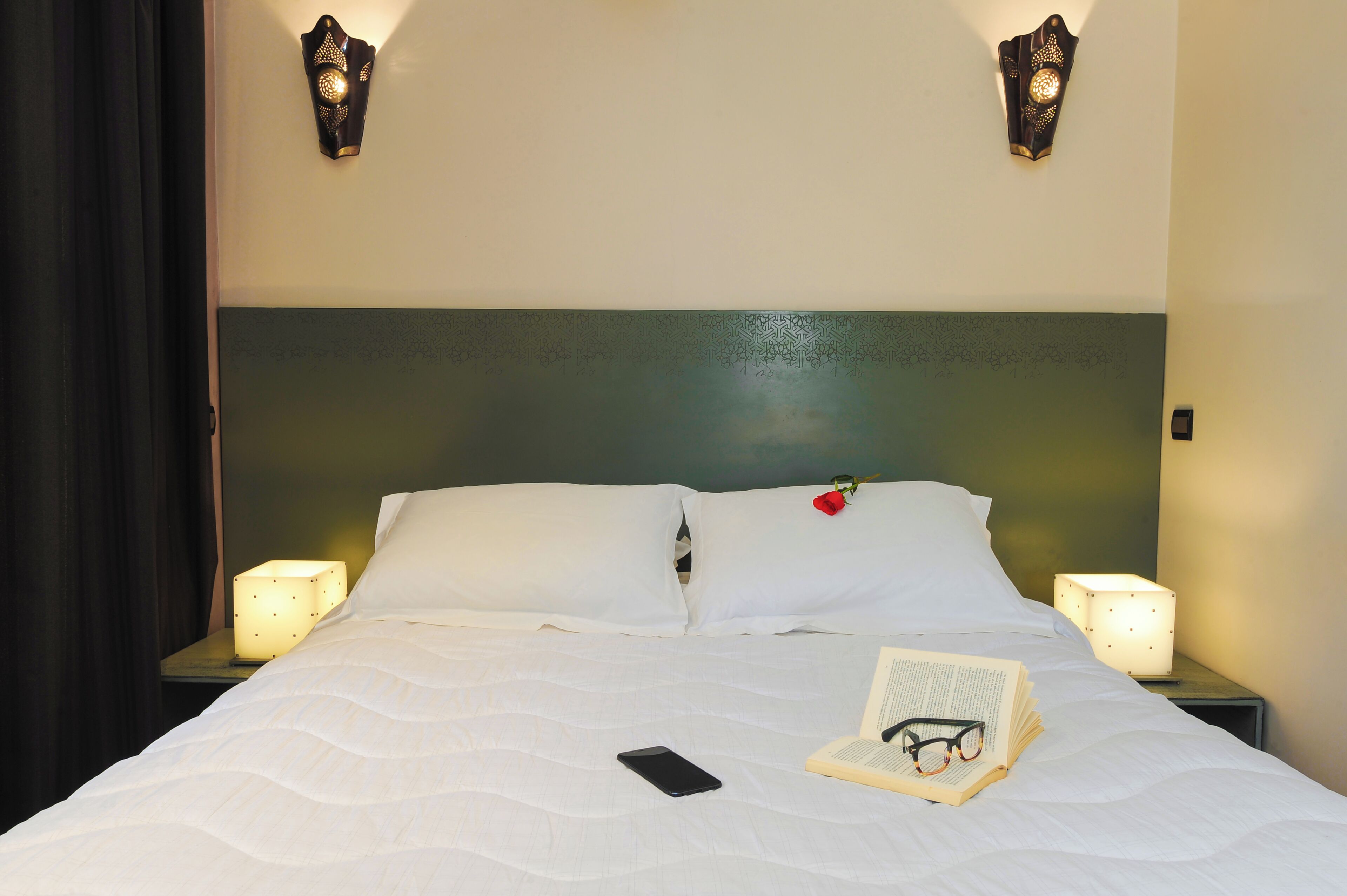 A neatly made bed with white linens, a rose on the pillow, and a book and glasses on the bed. There are two bedside lamps and a nightstand with a phone on it.