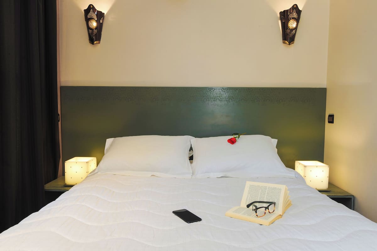 A neatly made bed with white linens, a rose on the pillow, and a book and glasses on the bed. There are two bedside lamps and a smartphone on the bed.