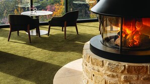 Una chimenea de piedra con frente de vidrio, una mesa redonda, dos sillas y vistas al paisaje a través de amplios ventanales.