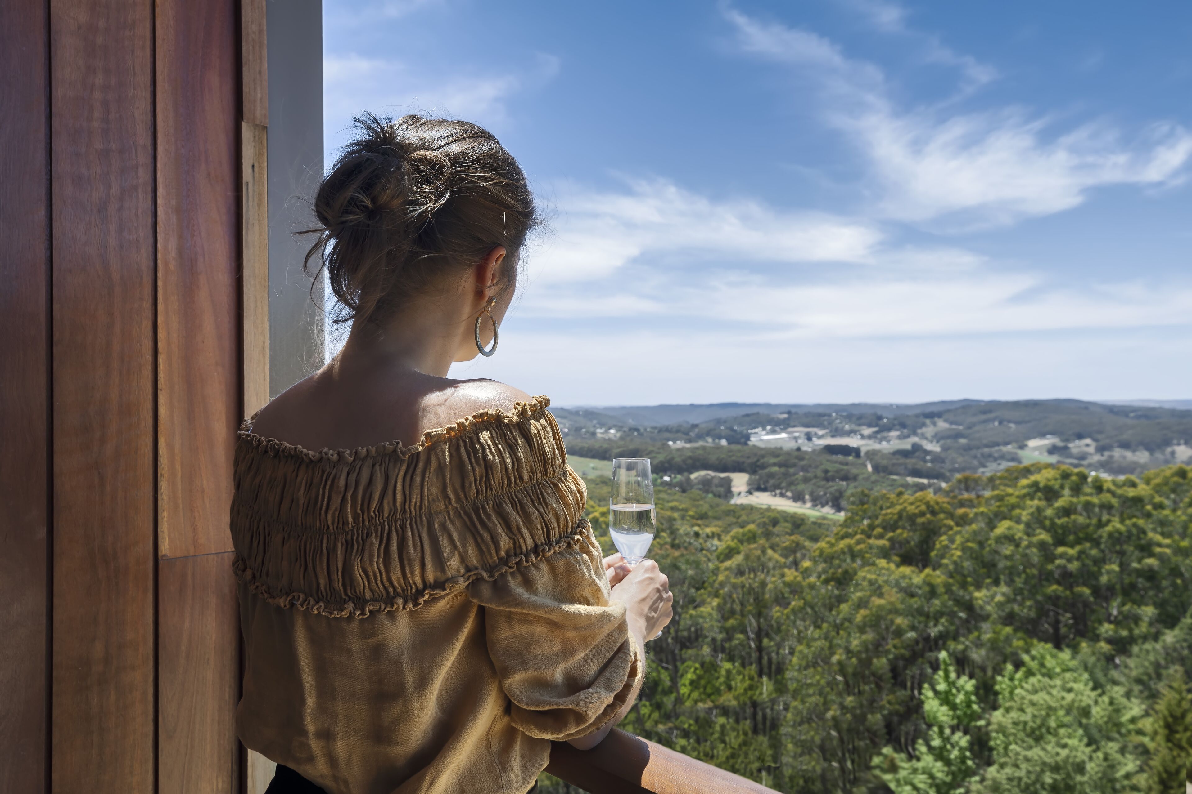 Iemand houdt een glas water vast en kijkt uit op een bebost landschap vanaf een balkon met houten leuningen.