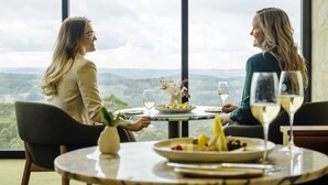 Dos personas cenando en una mesa con vistas panorámicas, un jarrón con flores y una copa de vino sobre la mesa.