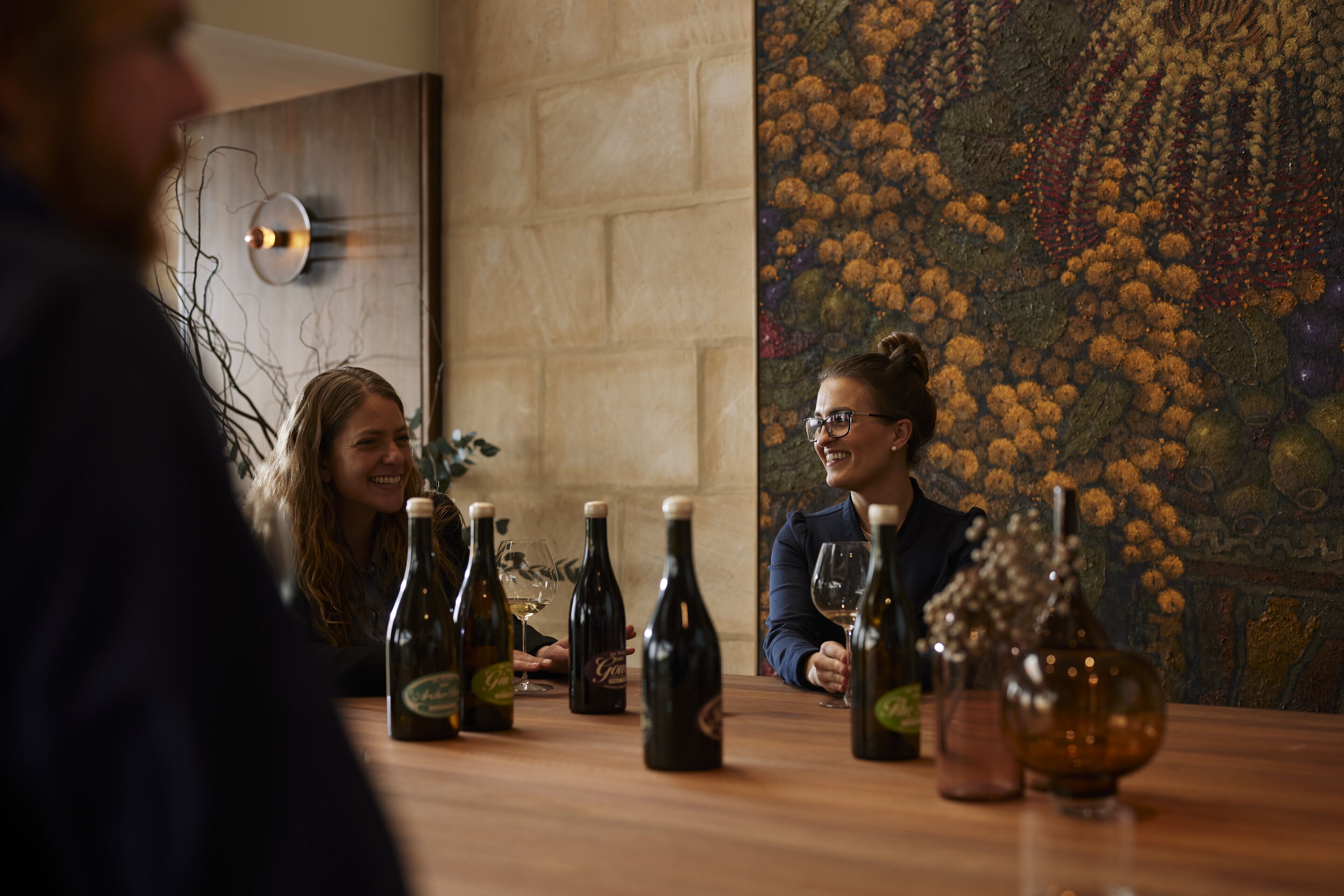Un grupo de personas sentadas alrededor de una mesa, con botellas y copas de vino, en una habitación con pared texturizada y un cuadro floral.