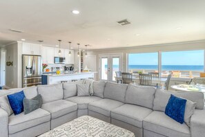 Una sala de estar moderna con un sofá modular grande, una cocina con armarios blancos y un comedor con vista al océano.
