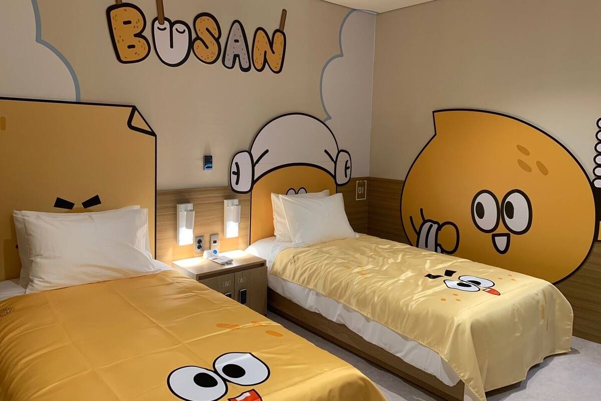 A hotel room with two beds, each with a cartoon character design on the bedspread. The room has a wall with a large cartoon character and the word "BUSAN" above the beds.