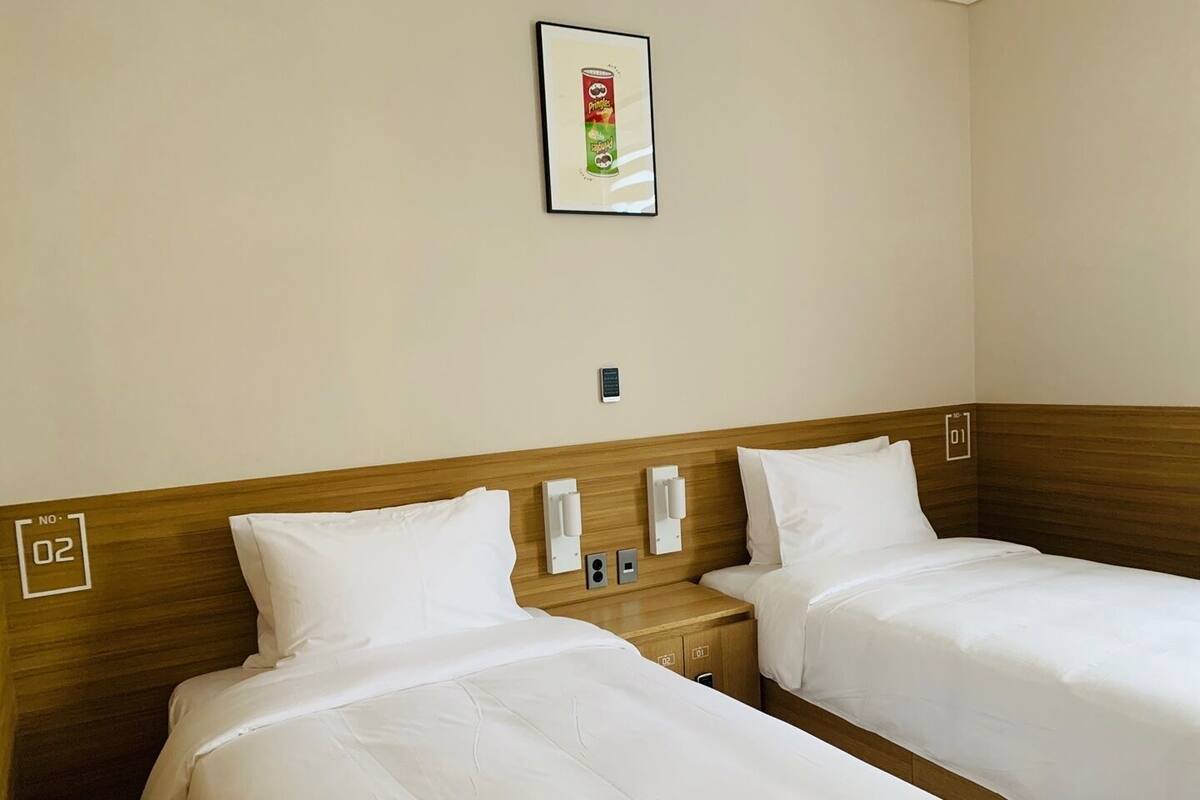 A hotel room with two single beds, wooden headboards, white bedding, a framed picture on the wall, and a small bedside table with a phone and a light switch.