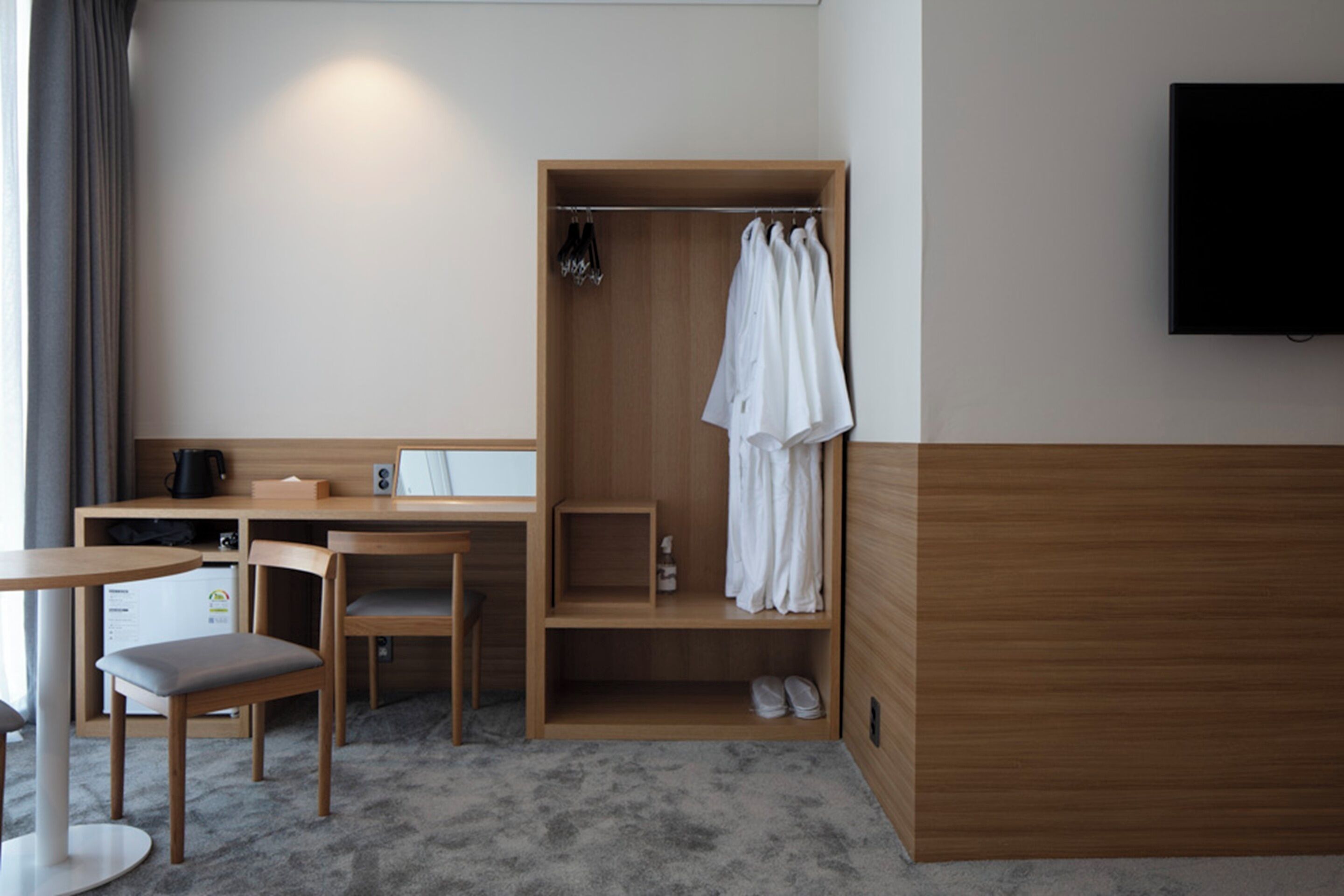 A hotel room with a wooden desk, a chair, a coat rack with a white robe, a television, and a shoe shelf.