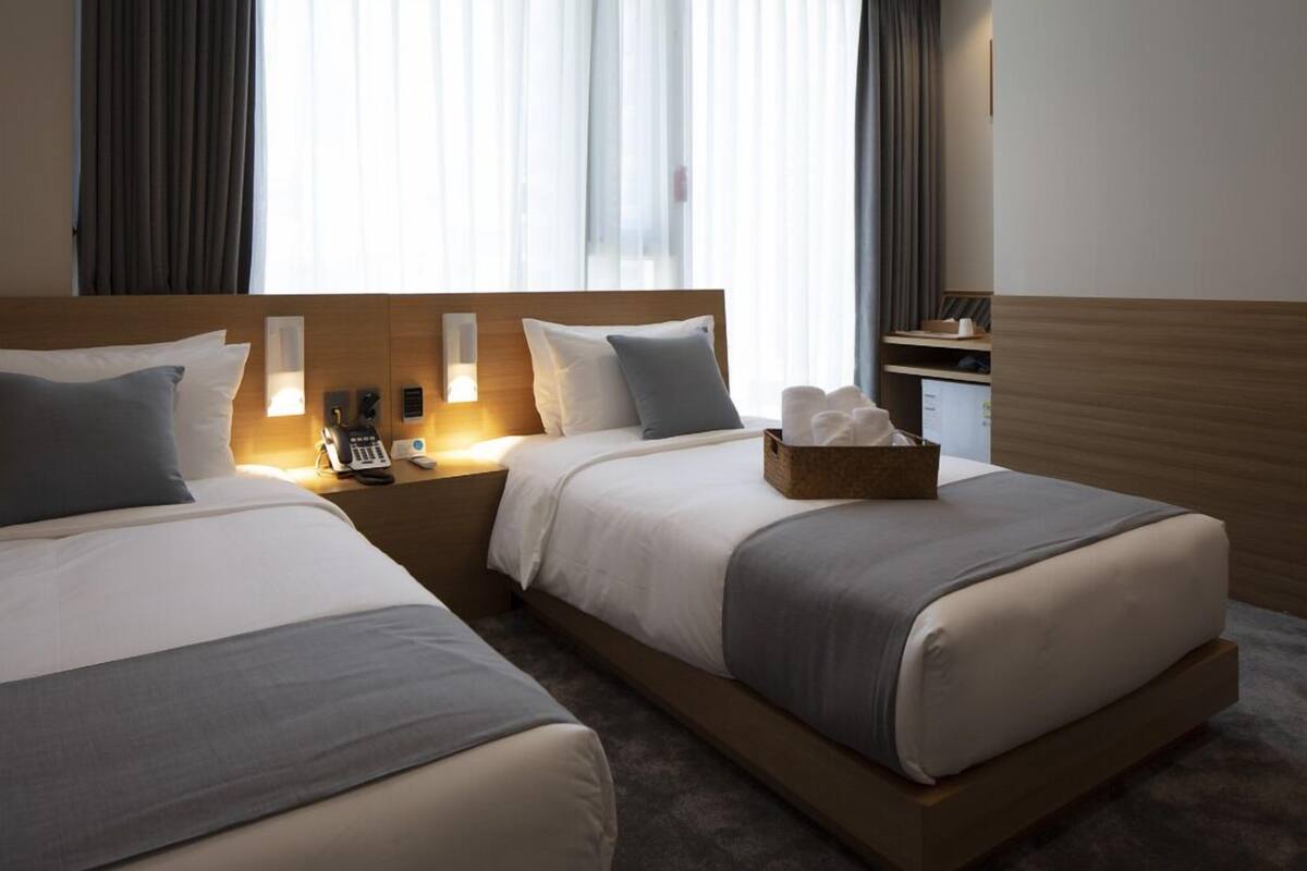 A hotel room with two beds, a wooden headboard, bedside lamps, a nightstand, a basket of toiletries, and a window with curtains.