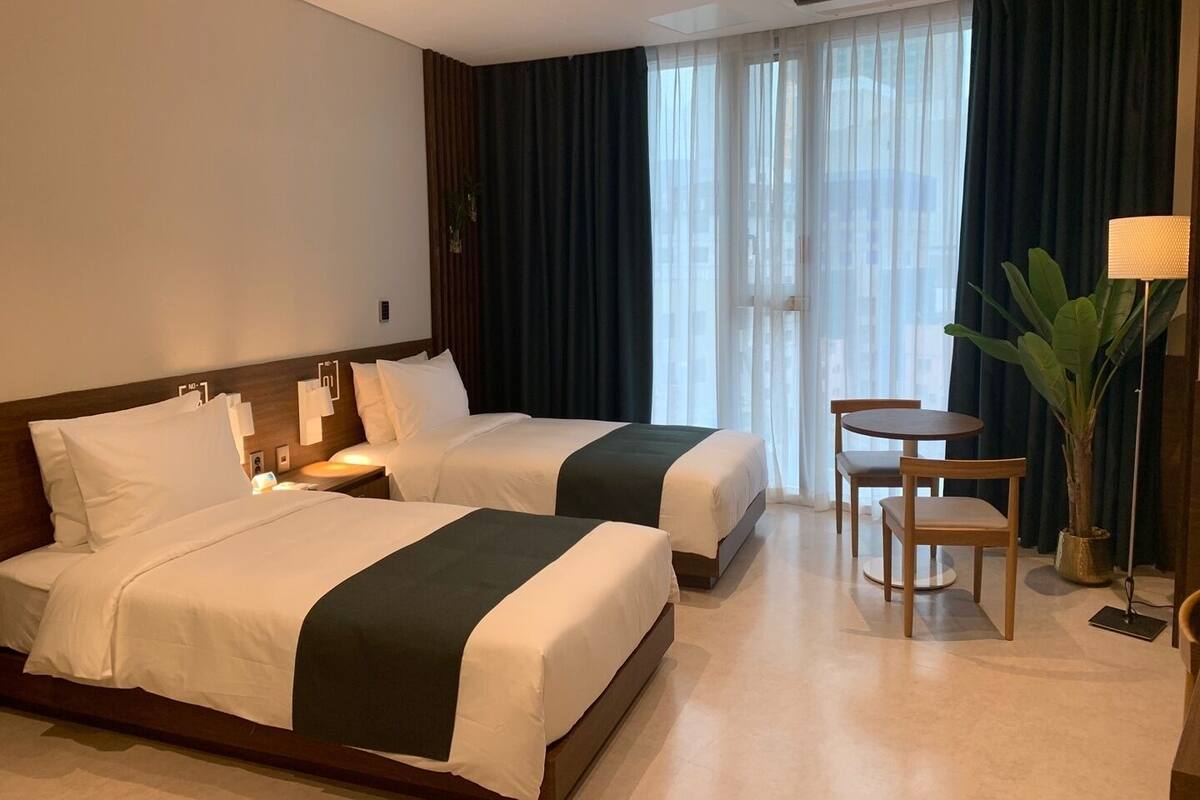A hotel room with two beds, a small table, a chair, and a large window with curtains.