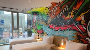 Una habitació d'hotel amb dos llits, un mural gran a la paret i vista a la ciutat a través de grans finestres.