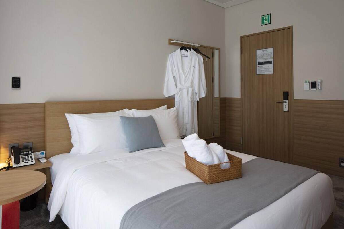 A hotel room with a bed, a bedside table with a phone, a robe hanging on a rack, and a basket of towels.