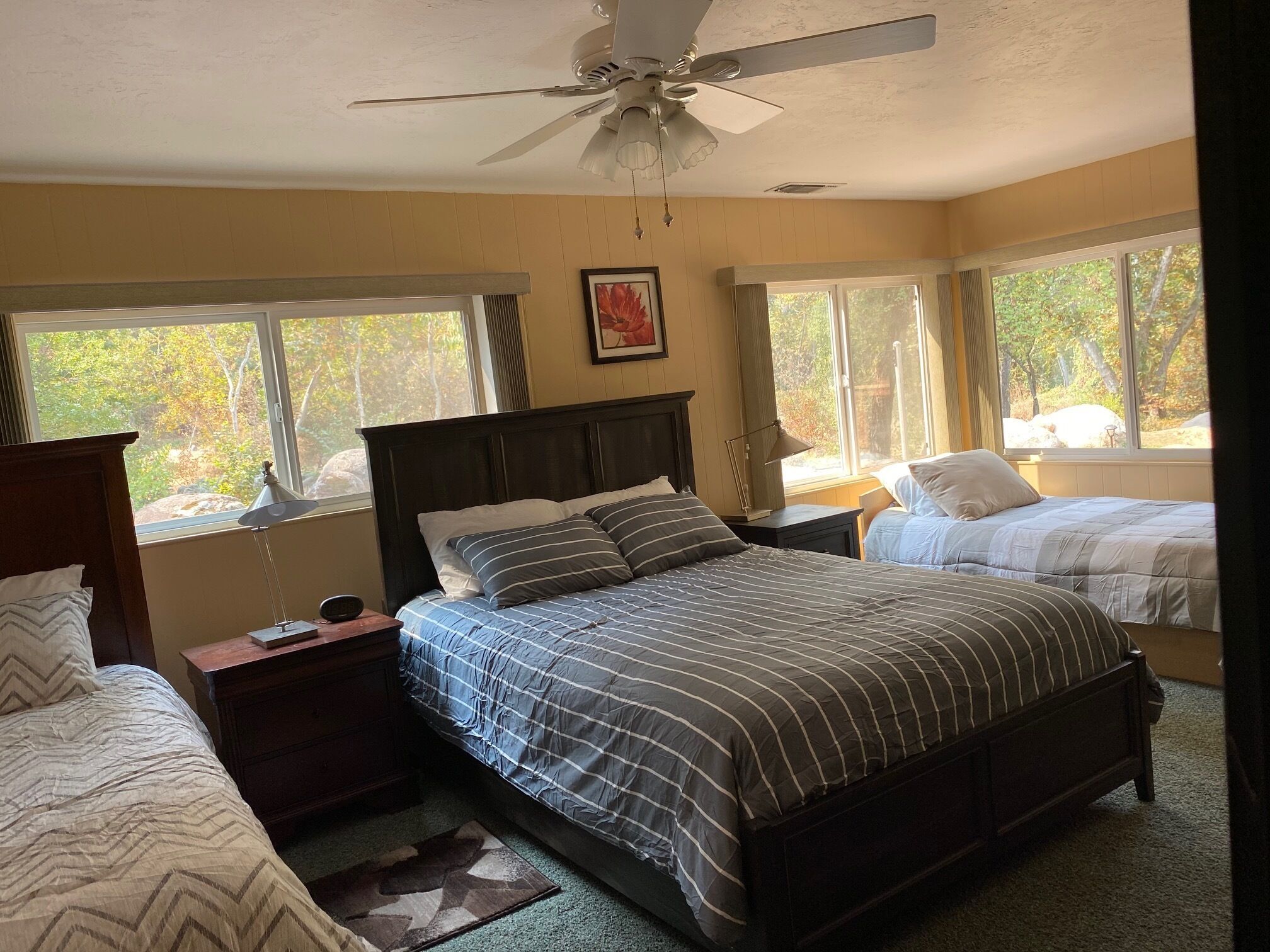 A bedroom with a large bed, two bedside tables, a ceiling fan, and windows with a view of trees.