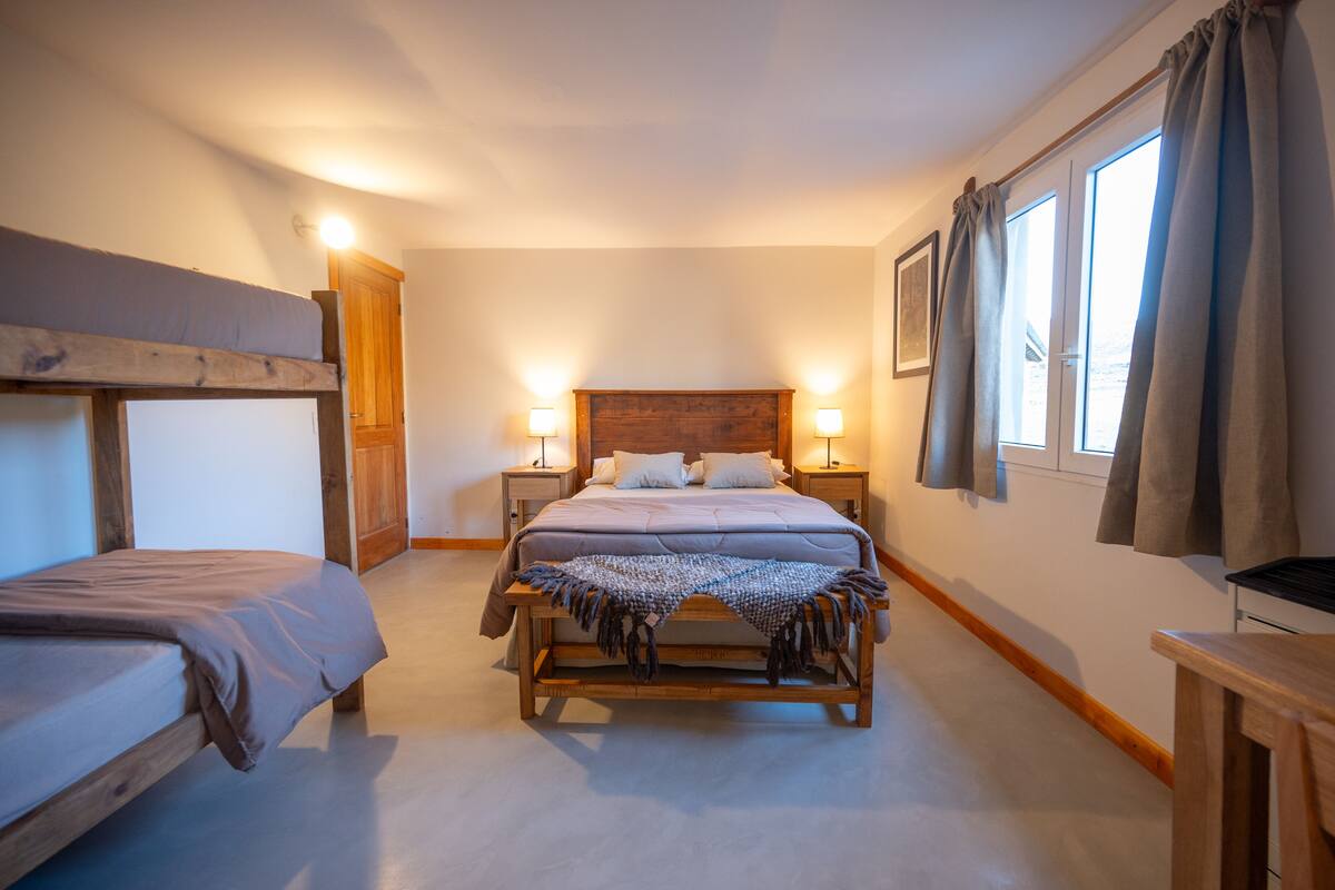 A bedroom with a large bed, a smaller bed, bedside lamps, a wooden headboard, and a window with curtains.