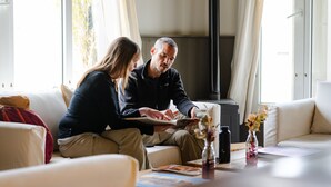 A couple sitting on a sofa with a book, a vase with flowers, and a fireplace in the background.