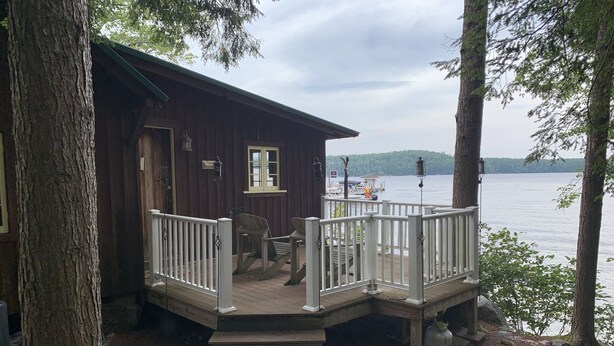 A wooden cabin with a deck overlooking a lake, featuring a small porch with chairs and a table. - Cozy Camp on Lake Winni (Meredith)