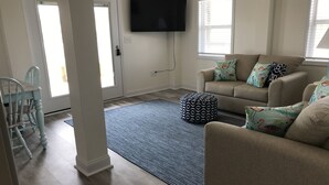 Ein Wohnzimmer mit einem beigen Sofa, gemusterten Kissen, einem blauen Teppich und einem Flachbildfernseher an der Wand.