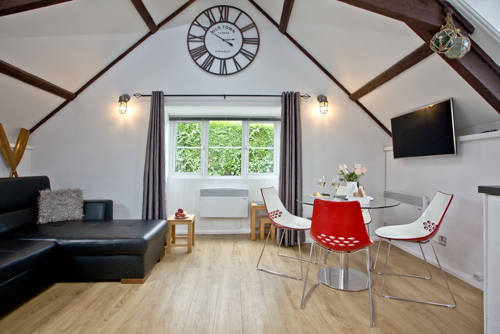 Une pièce confortable de style loft, avec un canapé en cuir noir, une table à manger entourée de chaises rouges et blanches, une grande horloge murale et une fenêtre agrémentée de rideaux.