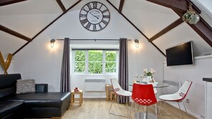 A cozy loft-style room with a black leather sofa, a dining table with red and white chairs, a large wall clock, and a window with curtains. - Free Parking - Free Wi-Fi - 2km From The Beach (Strete)