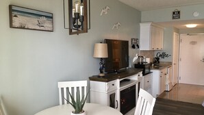 Un coin repas avec une table ronde, des chaises, une plante en pot, un luminaire fixé au mur et, au fond, une cuisine.