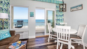 Une pièce avec vue sur l’océan, un coin repas avec des chaises blanches, un canapé marron agrémenté d’un coussin sur lequel est écrit « Beach Please », et une table avec des magazines.