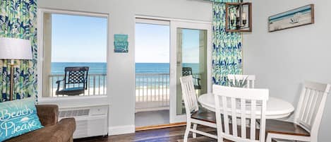 Quarto com vista para o oceano, área de jantar com cadeiras brancas, sofá marrom com almofada 'Beach Please' e uma mesa com revistas.