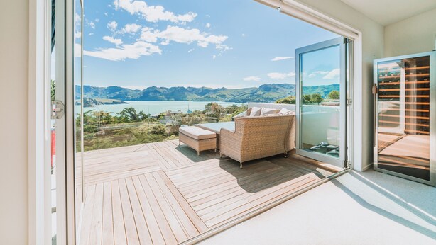 Een balkon met een rieten bank en een houten terras, met uitzicht op een meer en bergen.
