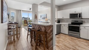 A modern kitchen with white cabinets, a stainless steel microwave, and an oven. There is a wooden bar stool and a window with a view of the outdoors. - Destin West Gulfside 510 ~ Amazing Beach Views ~ Beach Service (Fort Walton Beach)