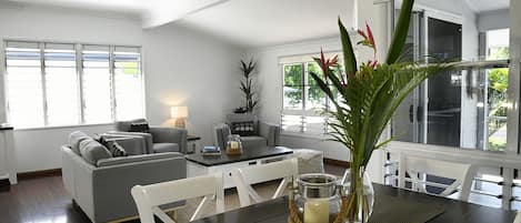 Una sala moderna con un sofá gris, una mesa de comedor con sillas y un ventanal con persianas.