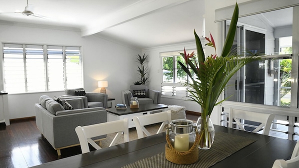 Un salon moderne avec un canapé gris, une table à manger avec des chaises et une grande fenêtre équipée de stores.